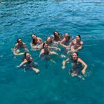 A group of people swimming and smiling in clear turquoise water, posing together in a sunny sea.