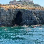 Turquoise sea by a rocky coastline with a dark sea cave and swimmers exploring near the entrance; a historic fortress sits atop the cliff above.