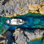 Aerial view of a white sailboat nestled in a narrow turquoise waterway between rocky cliffs.