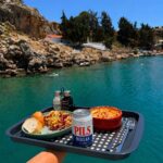 Tray with pasta, salad, bread, and a Pils Hellas beer can held over turquoise water and rocky coastline.